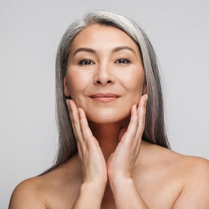 Beauty portrait of an attractive sensual mature topless woman with long gray hair standing isolated over gray background, posing, looking at camera